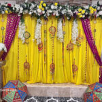 simple haldi decoration with a yellow wall backdrop, artificial flowers, colorful umbrellas, round hangings, decorative bells, and channel flowers