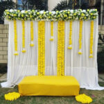 haldi decoration with white curtains backdrop long and short artificial string marigold flowers