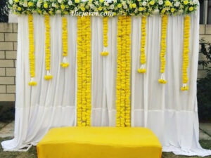 haldi decoration with white curtains backdrop long and short artificial string marigold flowers