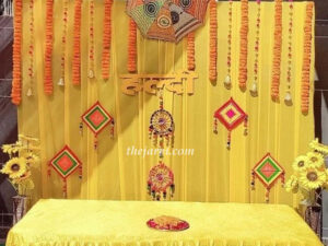 easy haldi decoration with orange backdrop, colorful umbrellas, orange marigold flowers (long & small mix), colorful round hangings, small bells and sitting table covered with cloth